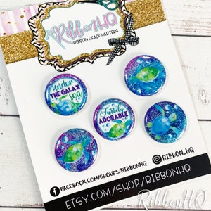 May include: Four 1.5 inch round resin buttons with a sea turtle theme. The buttons feature a blue and green turtle with a purple and blue glitter background. The text on the buttons reads "Under the Galax Sea", "Turtly Adorable", and two buttons with a turtle.