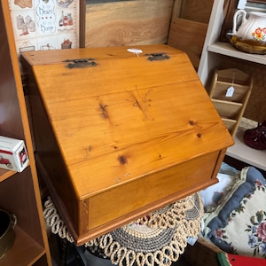May include: A wooden slant-top desk box with a natural honey-coloured finish, featuring a hinged lid with two dark metal hinges and a small, delicate floral engraving centred on the front panel, showcasing visible wood grain and knots.