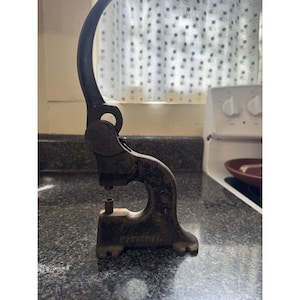 May include: An antique, rusty metal button maker with a curved handle. The base of the machine has the word "NATIONAL" stamped on it. The button maker is sitting on a speckled countertop.