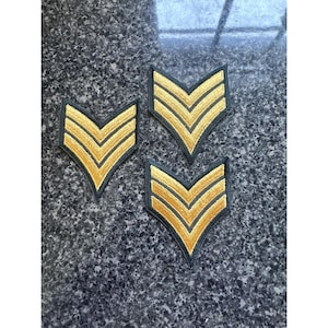 May include: Three gold and black chevron patches arranged on a dark, textured surface. Each patch features three gold stripes within a black border, forming a V-shape. The patches are likely for military or uniform decoration.