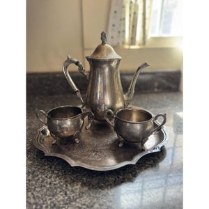 May include: A vintage silver tea set, including a teapot, two small handled cups, and a serving tray. The set has ornate detailing and is displayed on a speckled countertop. The teapot has a tall, slender design with a decorative lid.