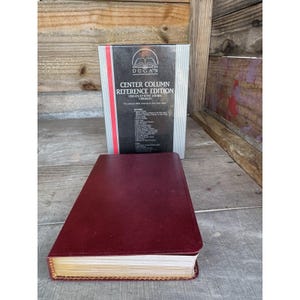 Op de afbeelding: Een bordeauxrood leren boek met goudgerande pagina's en een bijpassende doos. De doos bevat de tekst "DUGAN CENTER COLUMN REFERENCE EDITION" en een lijst met kenmerken. Het boek is open en toont de pagina's.