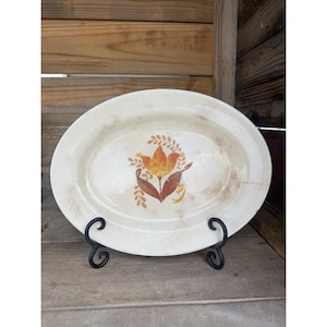 Könnte beinhalten: Ovale, cremefarbene Servierplatte mit einem Blumendesign in der Mitte. Die Blume ist gelb und orange mit braunen Blättern. Die Platte wird auf einem schwarzen Metallständer präsentiert, was einen Vintage-Look erzeugt.