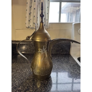 May include: A vintage brass coffee pot with a long spout and a decorative handle. The pot features etched designs of palm trees and camels. The lid is hinged and topped with a dark wooden finial. The pot is likely used for serving coffee or tea.