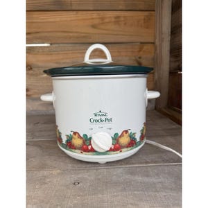Puede incluir: Olla de cocción lenta Rival Crock-Pot blanca con tapa y asa verde oscuro. La parte frontal del aparato tiene las palabras "Rival Crock-Pot" y un borde decorativo de peras y bayas. El mando tiene ajustes de Apagado, Bajo y Alto.
