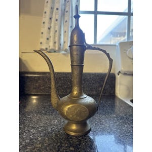 May include: A vintage brass pitcher with an elongated spout and ornate floral engravings. The pitcher has a tall, slender body with a curved handle and a decorative lid. The brass has a warm, aged patina.