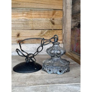 May include: An antique-style ceiling light fixture with a black chain and a decorative, ornate base. The base is a dark, metallic color with intricate detailing. A black, circular canopy is also visible.