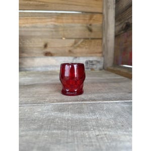 Op de afbeelding: Een klein, robijnrood glas met een geometrisch patroon. Het glas is cilindrisch met een licht uitlopende rand en staat op een houten oppervlak. De achtergrond is een houten muur.