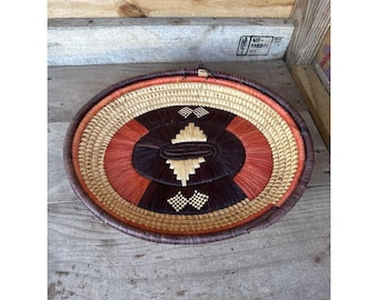 May include: A round woven basket with a decorative design. The basket features a central oval pattern in dark brown, surrounded by red-orange and natural tan hues. The rim is a darker brown.