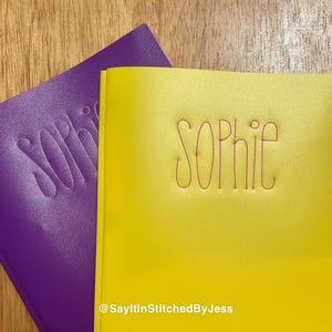 May include: Two notebook covers, one purple and one yellow. The name "Sophie" is stitched in red thread on both covers. The yellow cover has the text "@SayItInStitchedByJess" at the bottom.