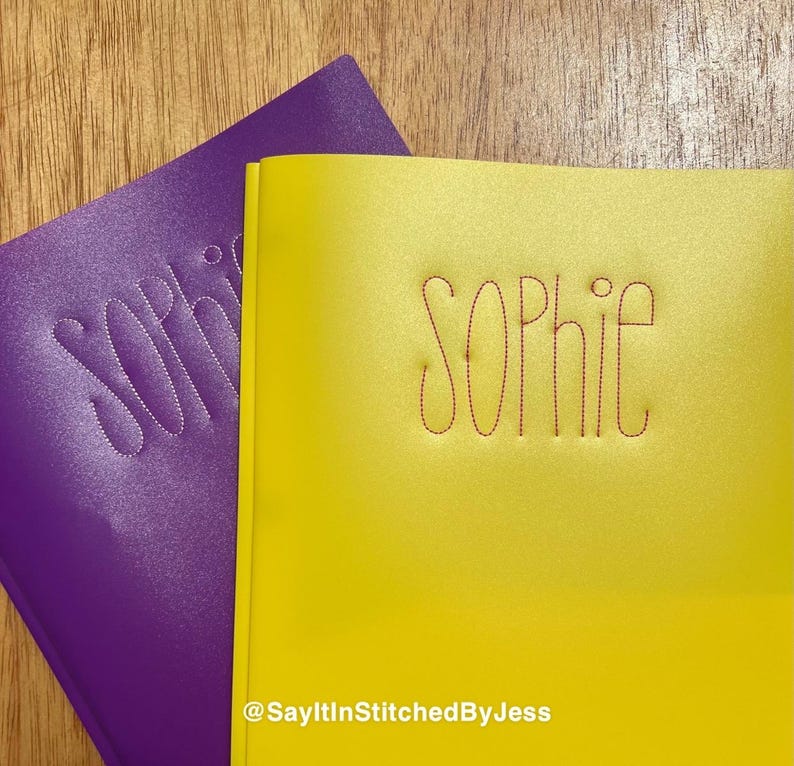 May include: Two notebook covers, one purple and one yellow. The name "Sophie" is stitched in red thread on both covers. The yellow cover has the text "@SayItInStitchedByJess" at the bottom.