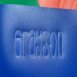 May include: Blue, green and red fabric with the name "Grayson" stitched in white thread. The fabric has a sparkly texture. The image includes the text "@SayItInStitchesByJess".