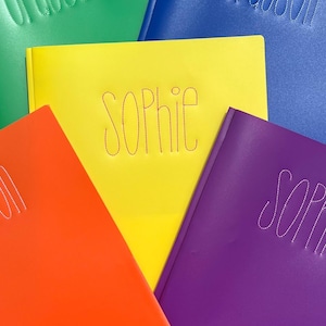 May include: Five colorful folders with names stitched on them. The folders are green, blue, yellow, orange, and purple. The names are Grayson, Sophie, and Grayson. The folders are on a wooden surface. The text at the bottom of the image reads "@SayItInStitchesByJess".