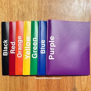 May include: A set of eight colourful binders in black, red, orange, yellow, green, blue, and purple. The binders are arranged in a row from left to right, with the black binder on the left and the purple binder on the right. The binders are all the same size and have a smooth, glossy finish.