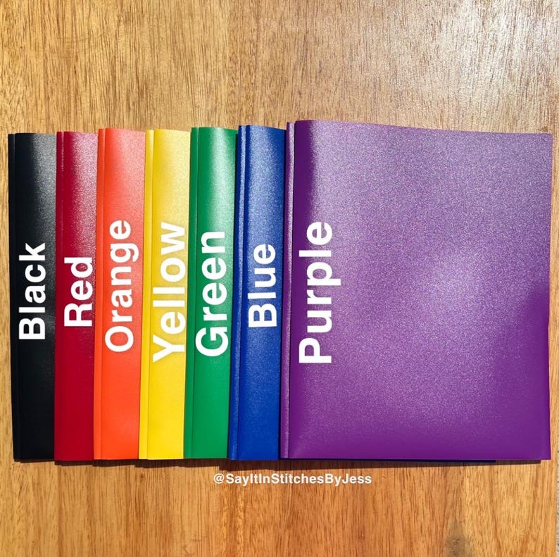 May include: A set of eight colourful binders in black, red, orange, yellow, green, blue, and purple. The binders are arranged in a row from left to right, with the black binder on the left and the purple binder on the right. The binders are all the same size and have a smooth, glossy finish.
