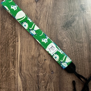 May include: A green and white camera strap with a white cat and flower pattern. The strap has a black adjustable strap and a small label that says "Crafty Husky".