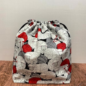 May include: A drawstring bag with a black base and a white fabric patterned with sheep and red yarn balls. The bag is designed to hold yarn or other craft supplies. The bag is sitting on a wooden surface.