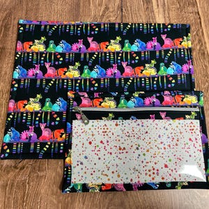 May include: Black fabric set with a colorful animal print. The set includes a rectangular mat and a zippered pouch with a clear window and a dotted pattern. The animal print features various colorful creatures on a black background.