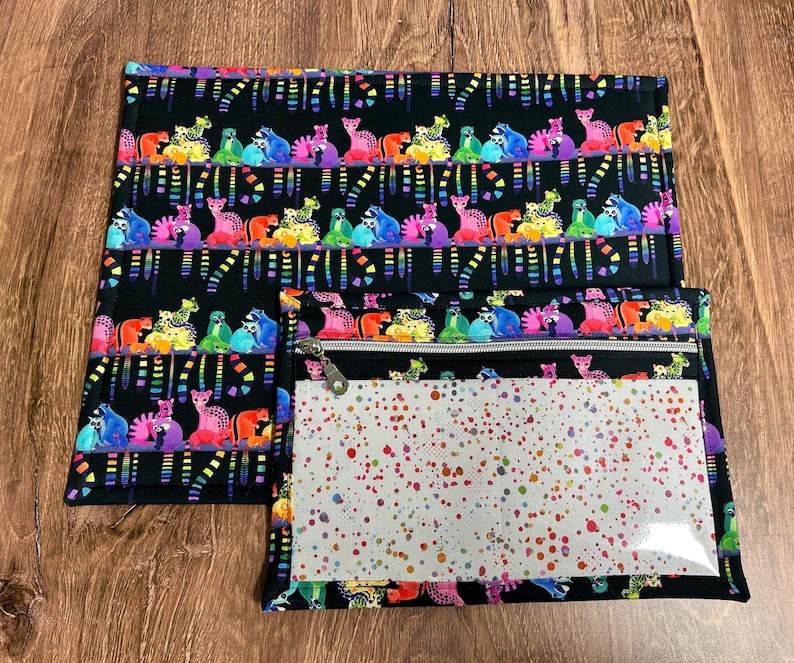 May include: Black fabric set with a colorful animal print. The set includes a rectangular mat and a zippered pouch with a clear window and a dotted pattern. The animal print features various colorful creatures on a black background.
