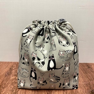 May include: A gray drawstring bag featuring a repeating pattern of various cat illustrations in black, white, and pink. The cats are in different poses, with some sleeping and others standing. The bag is made of fabric and has a drawstring closure.