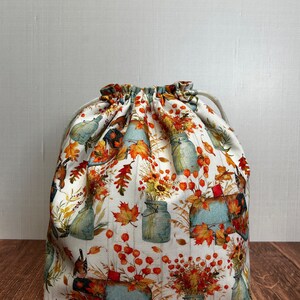 May include: A drawstring bag with a white background and a repeating pattern of autumn-themed illustrations. The design includes blue mason jars, sunflowers, and fall leaves in shades of orange, yellow, and brown. The bag is made of fabric and has a drawstring closure.