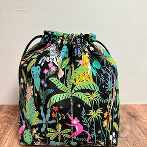 May include: A black drawstring bag with a vibrant jungle print. The fabric features colourful illustrations of zebras, leopards, monkeys, and lush foliage. The bag has a black drawstring closure and is set against a wooden surface.