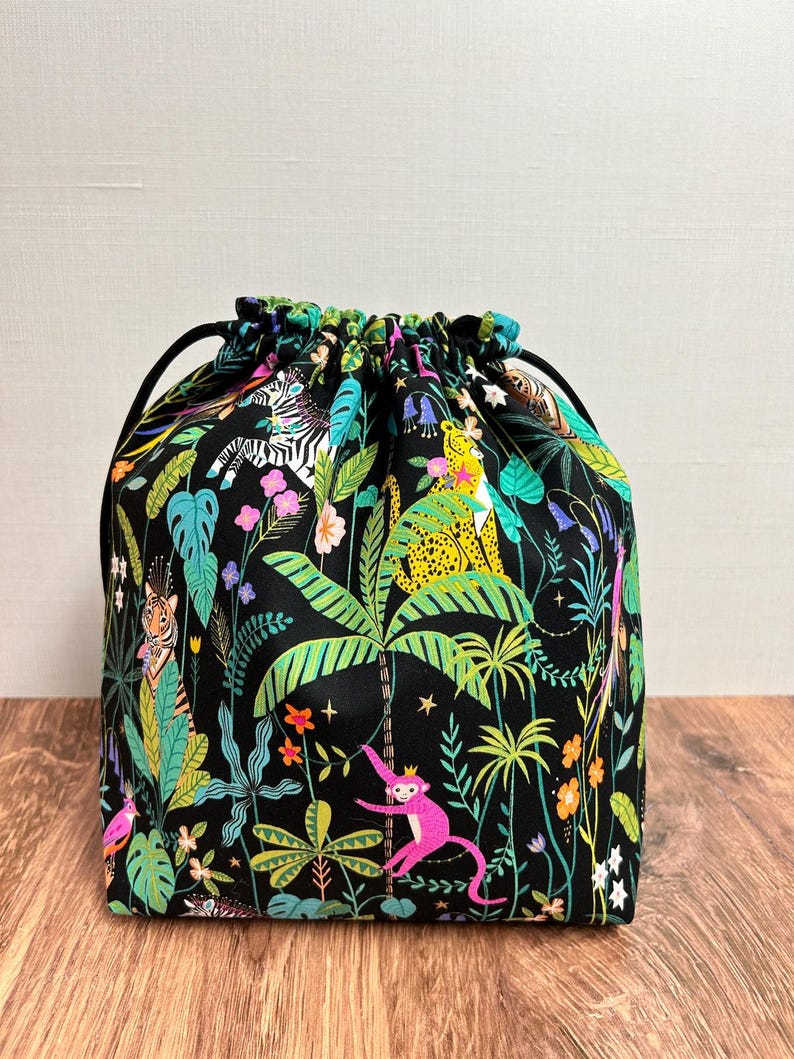 May include: A black drawstring bag with a vibrant jungle print. The fabric features colourful illustrations of zebras, leopards, monkeys, and lush foliage. The bag has a black drawstring closure and is set against a wooden surface.