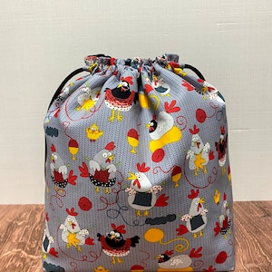 May include: A gray drawstring bag with a pattern of chickens knitting yarn balls. The chickens are red, yellow, and black with white accents. The background is a gray knitted pattern.