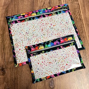 May include: Two clear zippered pouches with a white interior speckled with colorful paint splatters. The pouches are trimmed with black fabric and a colorful pattern of cartoon creatures. The zippers are silver.
