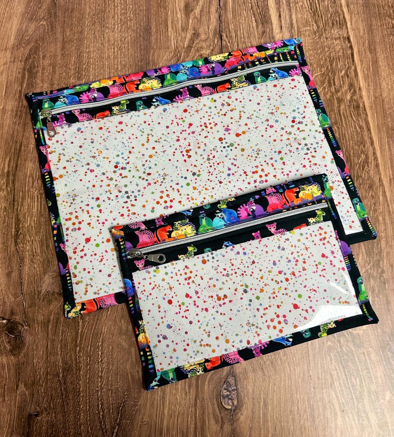 May include: Two clear zippered pouches with a white interior speckled with colorful paint splatters. The pouches are trimmed with black fabric and a colorful pattern of cartoon creatures. The zippers are silver.