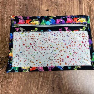 May include: A rectangular black zippered pouch with a clear front panel. The pouch features a colorful animal print border and a white interior with multicolored paint splatters. The zipper is silver.