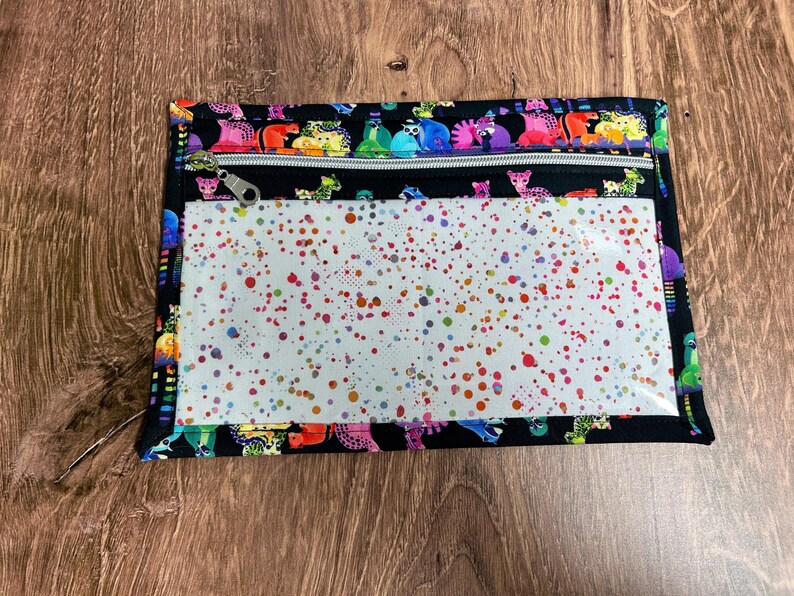 May include: A rectangular black zippered pouch with a clear front panel. The pouch features a colorful animal print border and a white interior with multicolored paint splatters. The zipper is silver.