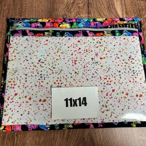 May include: A rectangular zippered pouch with a clear front and a colorful spotted pattern. The edges are trimmed with a black fabric featuring a repeating pattern of colorful cartoon animals. The pouch measures 11 x 14 inches.
