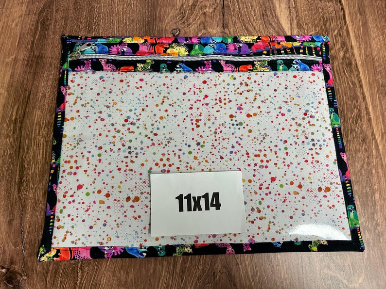 May include: A rectangular zippered pouch with a clear front and a colorful spotted pattern. The edges are trimmed with a black fabric featuring a repeating pattern of colorful cartoon animals. The pouch measures 11 x 14 inches.