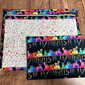 May include: Two fabric pouches with colorful designs. One pouch has a zipper and a white interior with colorful splatters. The other pouch is smaller and has a black background with colorful animal illustrations.