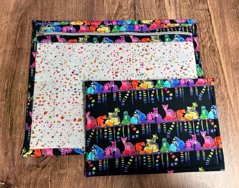 May include: Two fabric pouches with colorful designs. One pouch has a zipper and a white interior with colorful splatters. The other pouch is smaller and has a black background with colorful animal illustrations.