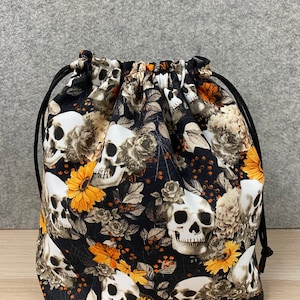 May include: A black drawstring bag with a skull and floral print. The print features white skulls, orange sunflowers, and brown leaves on a black background.