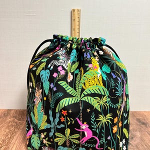 May include: A drawstring bag featuring a jungle design. The black fabric is adorned with vibrant illustrations of zebras, monkeys, tigers, and tropical plants. It has a black drawstring closure. The bag is on a wooden surface.