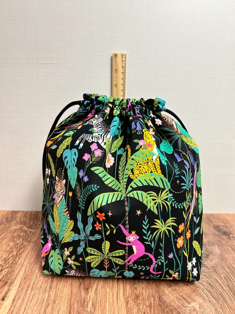 May include: A drawstring bag featuring a jungle design. The black fabric is adorned with vibrant illustrations of zebras, monkeys, tigers, and tropical plants. It has a black drawstring closure. The bag is on a wooden surface.