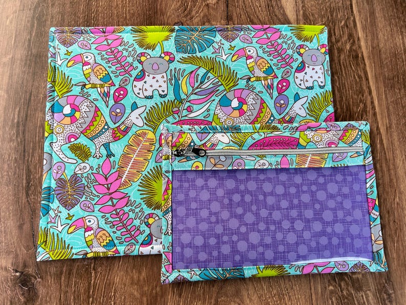 Puede incluir: Dos bolsas de tela con un colorido estampado tropical con tucanes, koalas y otros animales. La bolsa m&aacute;s grande es rectangular, y la m&aacute;s peque&ntilde;a tiene una ventana p&uacute;rpura transparente y una cremallera.