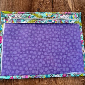 Puede incluir: Una bolsa rectangular con cremallera, con un panel frontal transparente y un estampado de lunares morados. La bolsa est&aacute; adornada con una colorida tela estampada con dise&ntilde;os caprichosos en tonos azules, rosas y amarillos.