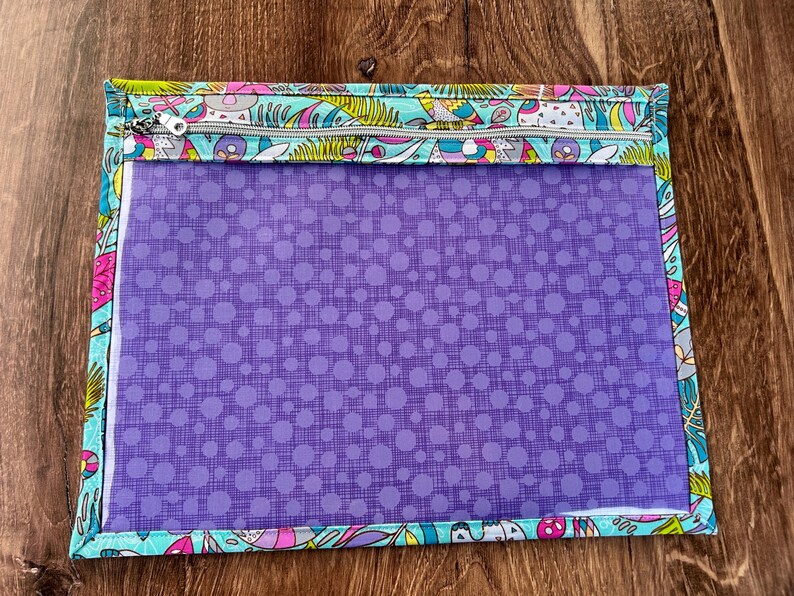 Puede incluir: Una bolsa rectangular con cremallera, con un panel frontal transparente y un estampado de lunares morados. La bolsa est&aacute; adornada con una colorida tela estampada con dise&ntilde;os caprichosos en tonos azules, rosas y amarillos.