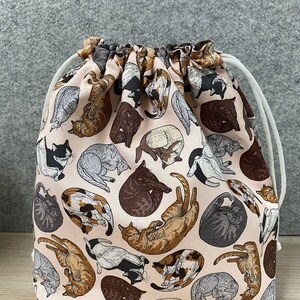 May include: A drawstring bag featuring a repeating pattern of various cat illustrations in shades of brown, gray, and black on a light pink background. The bag has a white drawstring closure.