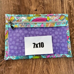 Puede incluir: Una bolsa rectangular con cremallera, con una parte delantera transparente y un borde de tela estampada. El borde presenta un dise&ntilde;o colorido con p&aacute;jaros y flores. La bolsa tiene un interior morado con una etiqueta blanca que dice "7x10".
