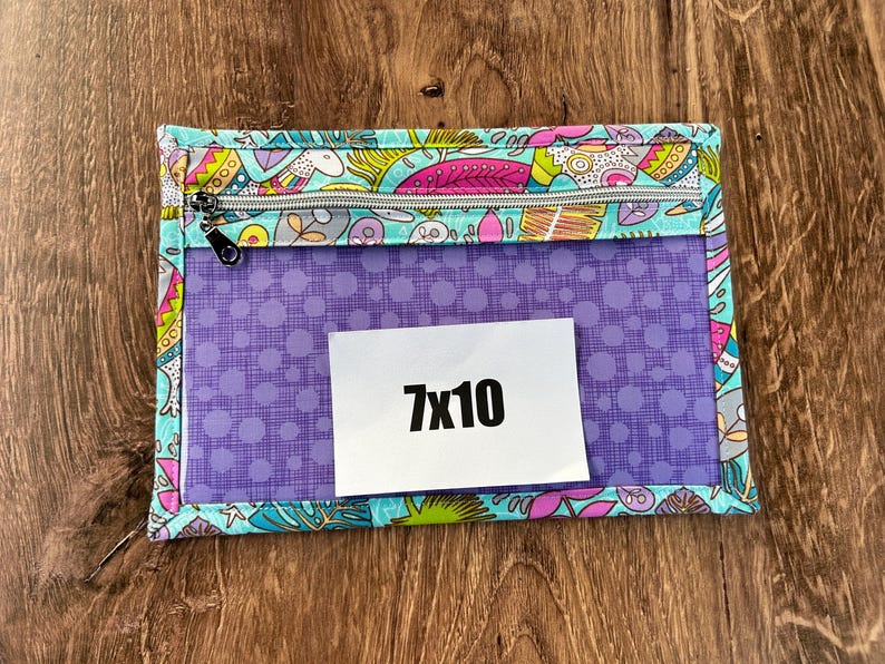 Puede incluir: Una bolsa rectangular con cremallera, con una parte delantera transparente y un borde de tela estampada. El borde presenta un dise&ntilde;o colorido con p&aacute;jaros y flores. La bolsa tiene un interior morado con una etiqueta blanca que dice "7x10".