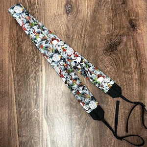 May include: A black camera strap with a white and colorful pattern of cartoon dogs. The strap has adjustable length and a metal clip.