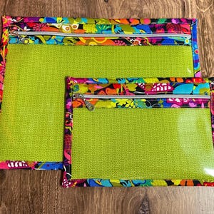 Puede incluir: Dos bolsas transparentes con cremallera, con un interior verde brillante y un borde con estampado floral colorido. Las bolsas son rectangulares, una m&aacute;s grande que la otra, y est&aacute;n sobre una superficie de madera.