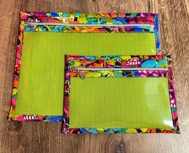 Puede incluir: Dos bolsas transparentes con cremallera, con un interior verde brillante y un borde con estampado floral colorido. Las bolsas son rectangulares, una m&aacute;s grande que la otra, y est&aacute;n sobre una superficie de madera.