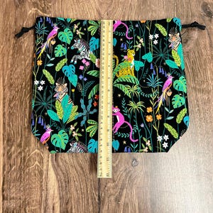 May include: Black drawstring bag with a vibrant jungle print featuring tigers, zebras, monkeys, and parrots amidst lush foliage. The bag measures approximately 30 cm tall. Black drawstrings secure the top.
