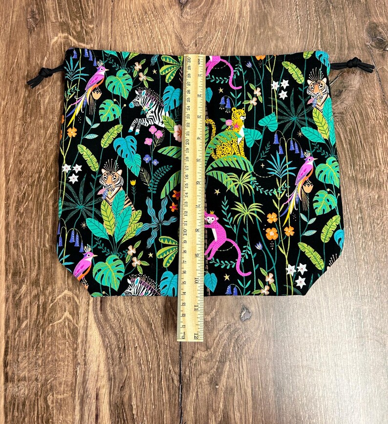 May include: Black drawstring bag with a vibrant jungle print featuring tigers, zebras, monkeys, and parrots amidst lush foliage. The bag measures approximately 30 cm tall. Black drawstrings secure the top.